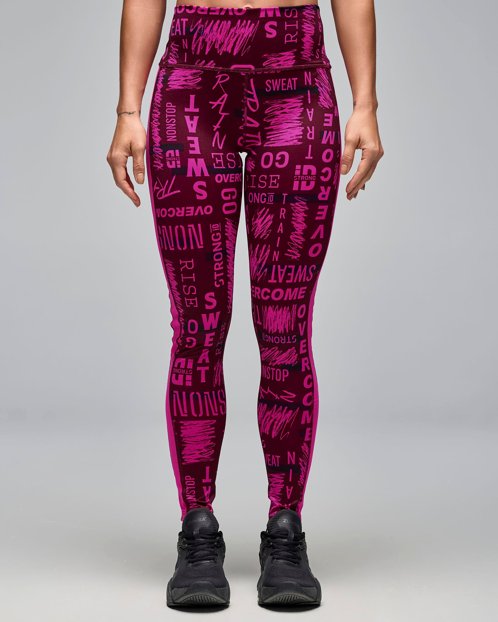 Burgundy and pink high-waisted ankle leggings with an allover motivational word print. Female model wears the fitted leggings with black athletic sneakers in a front view against a light gray background.