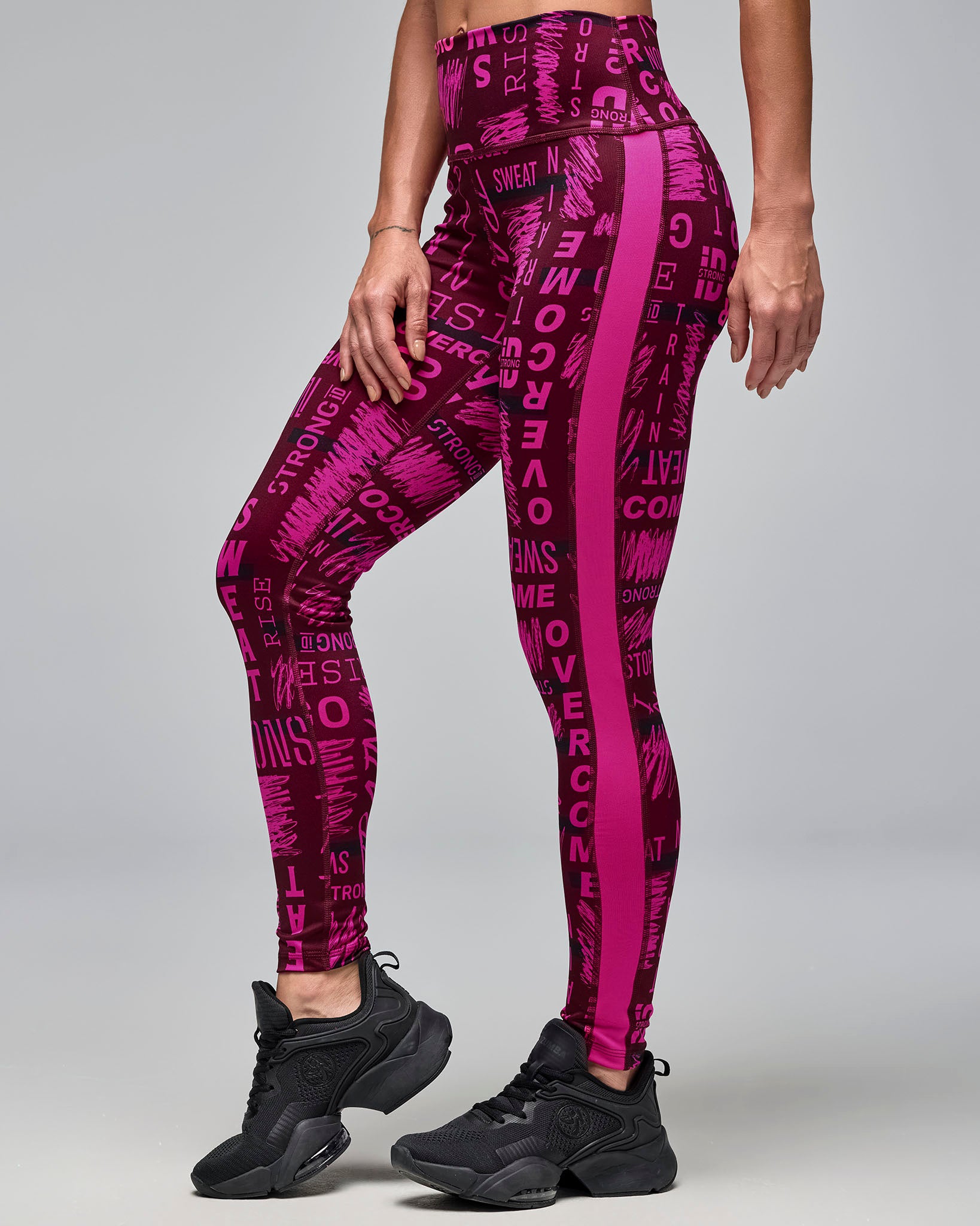 Burgundy and pink high-waisted ankle length leggings with an allover motivational word print and bright pink side panels. Female model wears the fitted leggings with black athletic sneakers in a side view against a light gray background.