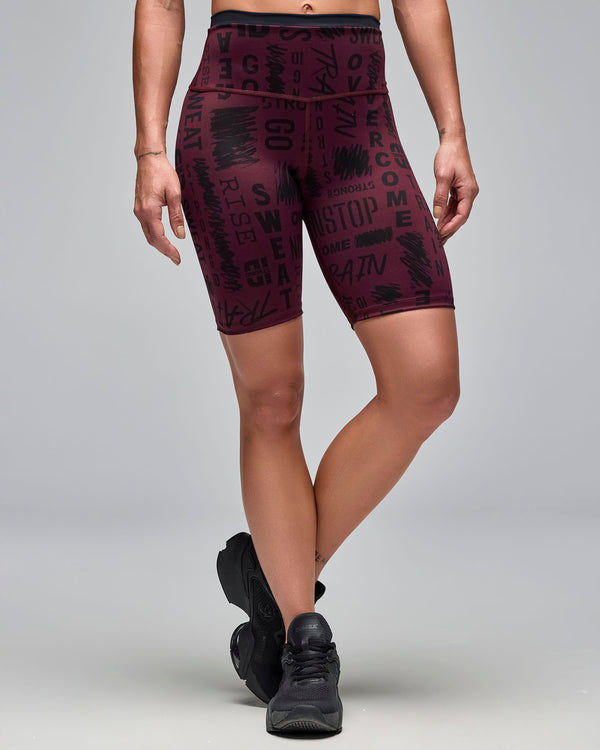 Burgundy high-waisted biker shorts with an allover black motivational word print. Female model wears the fitted mid-thigh shorts with black athletic sneakers in a front view against a light gray background.