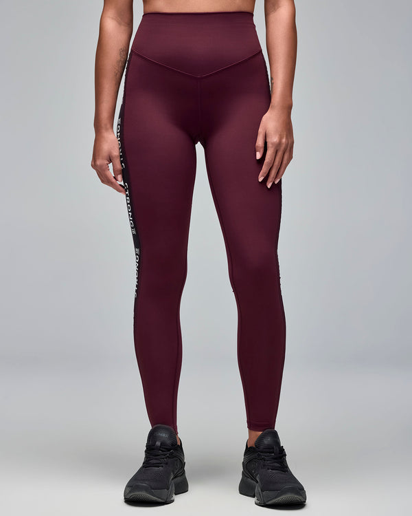 Burgundy high-waisted full-length leggings with a smooth fitted front and black side text panels. Female model wears the leggings with black athletic sneakers in a front view against a light gray background.