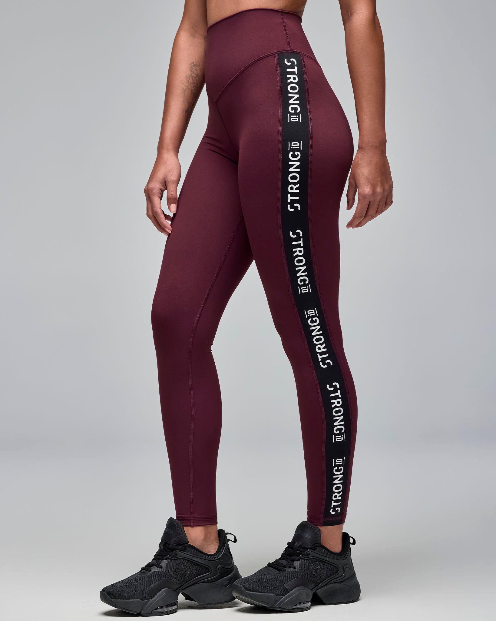 Burgundy high-waisted full-length leggings with black side panels featuring repeated white “STRONG iD” text. Female model wears the fitted leggings with black athletic sneakers in a side view against a light gray background.