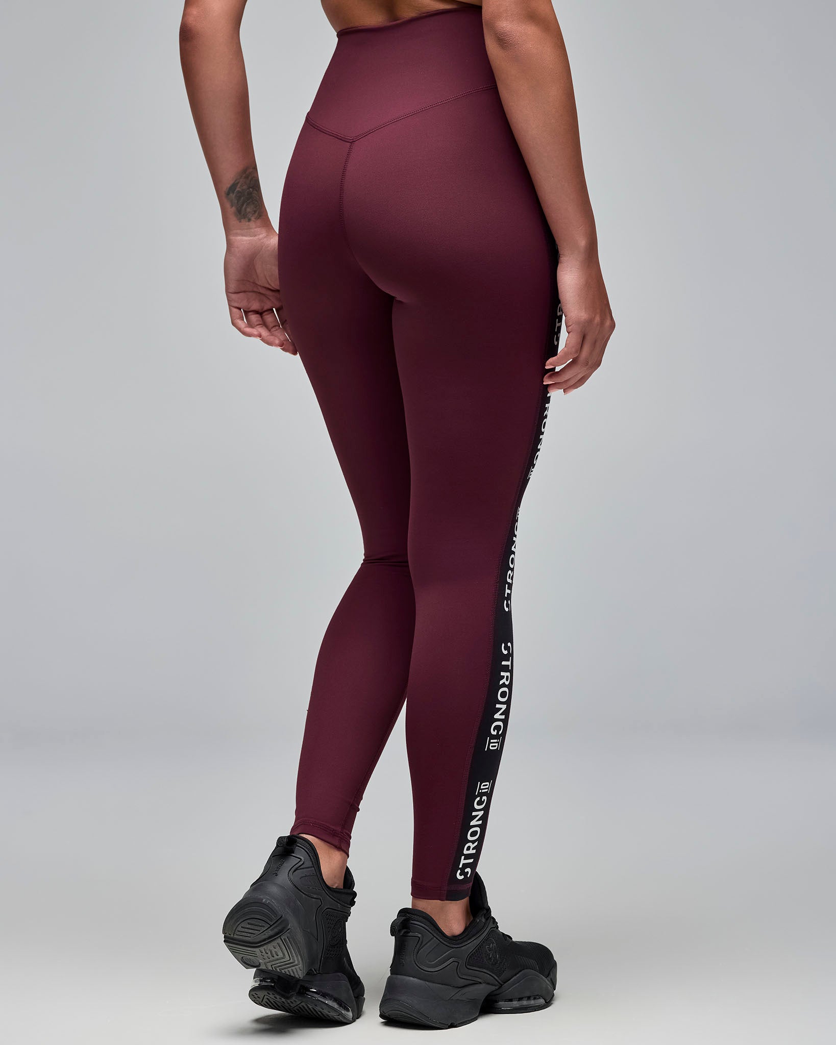 Back view of burgundy high-waisted full-length leggings with black side panels and repeated white “STRONG iD” text. Female model wears the fitted leggings with black athletic sneakers against a light gray background.