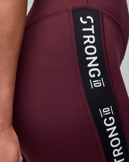 Burgundy high-waisted leggings with a black side panel and repeated white “STRONG iD” text. Female model wears the fitted full-length leggings in a close-up side view against a light gray background.