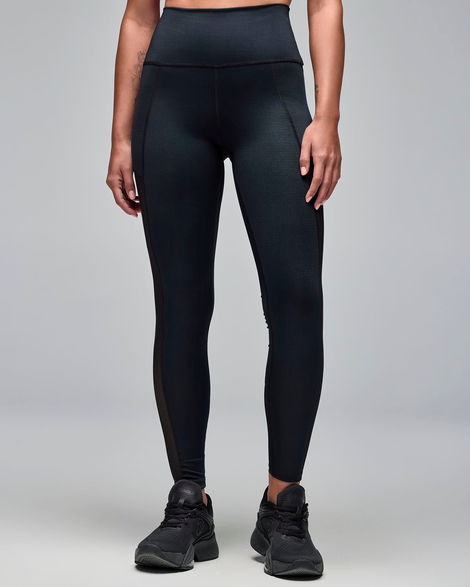 Black high-waisted full-length leggings with a smooth fitted silhouette. Female model wears the leggings with black athletic sneakers in a front view against a light gray background.