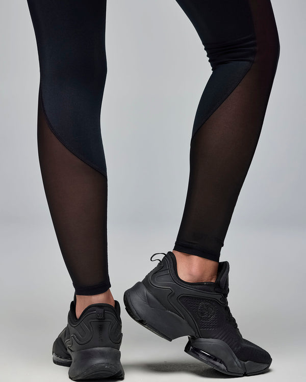 Black high-waisted leggings with sheer mesh side panels, paired with black athletic sneakers. Female model wears the fitted full-length leggings in a lower-leg view against a light gray background.