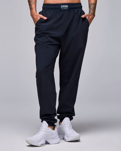 Female model in the black high-waisted joggers, with a closer look at the waistband label: white 'STRONG' text over the tagline 'UNITED IN OUR QUEST TO BE UNSTOPPABLE.'