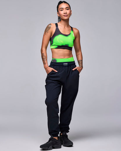 Full view of a female model in the black high-waisted joggers, paired with a neon green and black swirl print bra and black shoes.