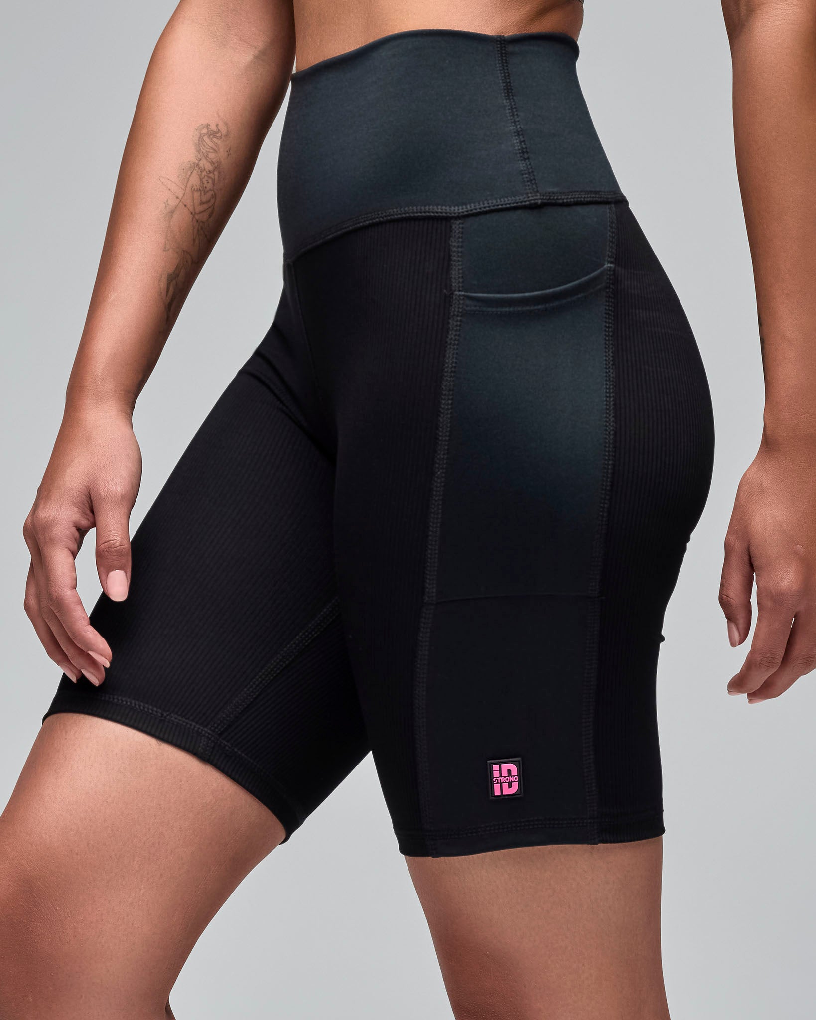 Black high-waisted biker shorts with ribbed texture panels, side pockets, and a small pink "STRONG iD" logo near the hem. Female model wears the fitted mid-thigh shorts in a side view against a light gray background.