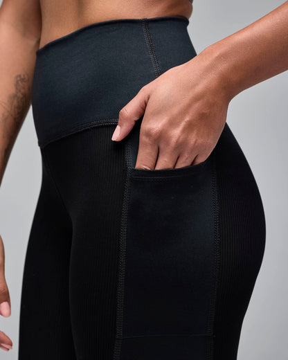 Female model wears the fitted mid-thigh shorts in a close-up view showing a hand in the pocket against a light gray background. She wears a black high-waisted biker shorts with ribbed texture panels and a side pocket. 