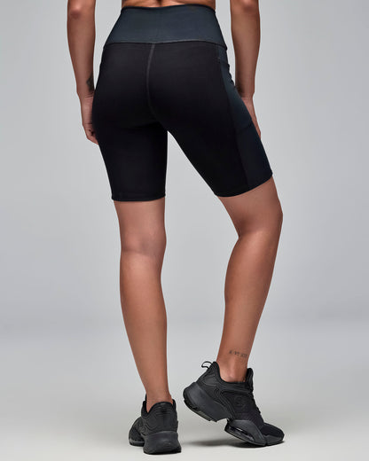 Back view of black high-waisted biker shorts with ribbed texture panels and side pockets. Female model wears the fitted mid-thigh shorts with black athletic sneakers against a light gray background.