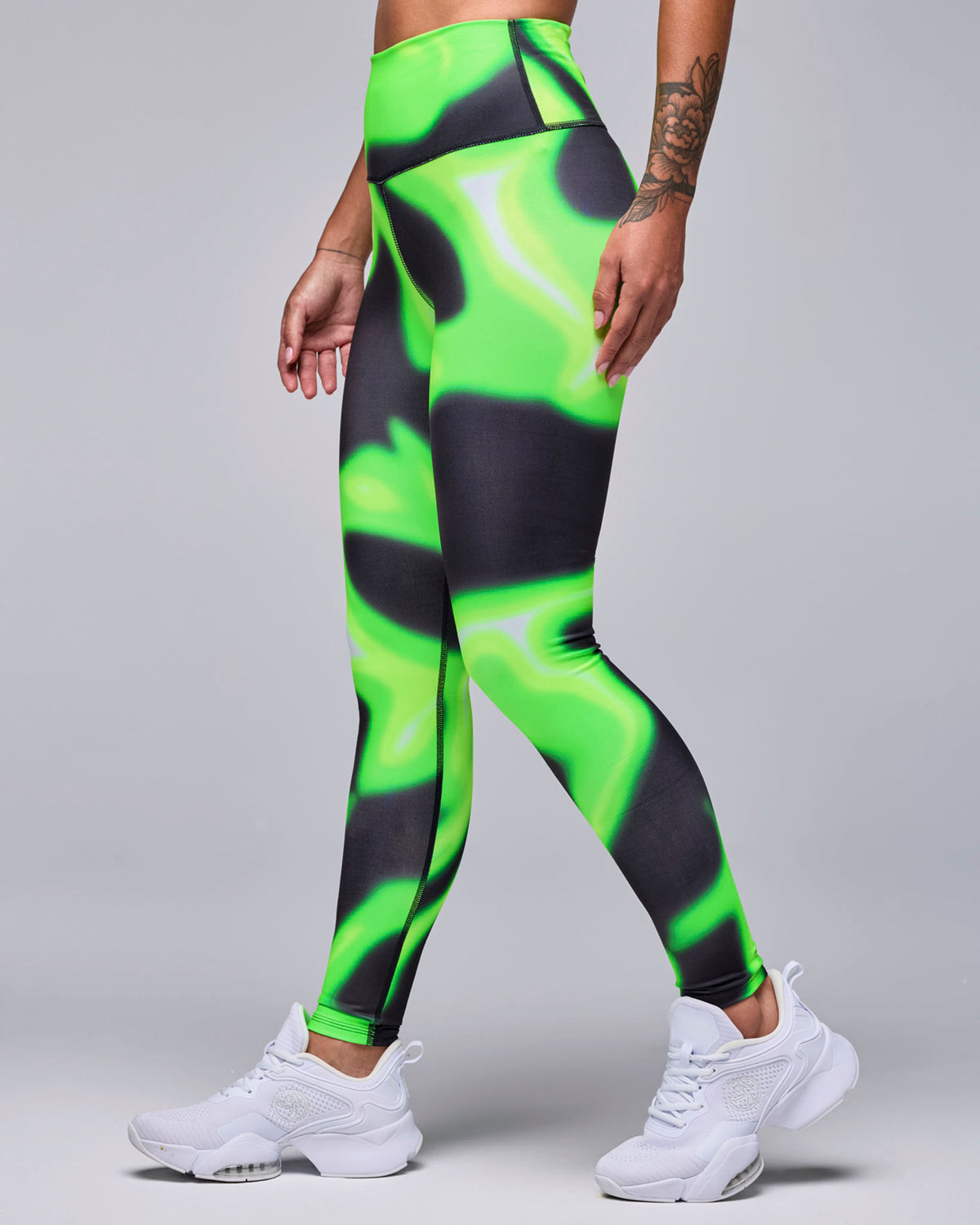 Female model in high-waisted ankle leggings with a neon green and black swirl print and a smooth, wide waistband, shown in side view with white shoes.
