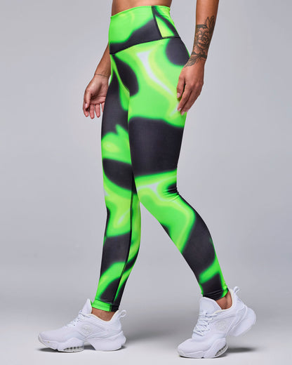 Female model in high-waisted ankle leggings with a neon green and black swirl print and a smooth, wide waistband, shown in side view with white shoes.