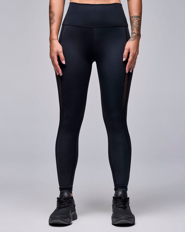 Black high-waisted ankle leggings on a female model, with a wide, smooth waistband and mesh inserts visible along both outer thighs.