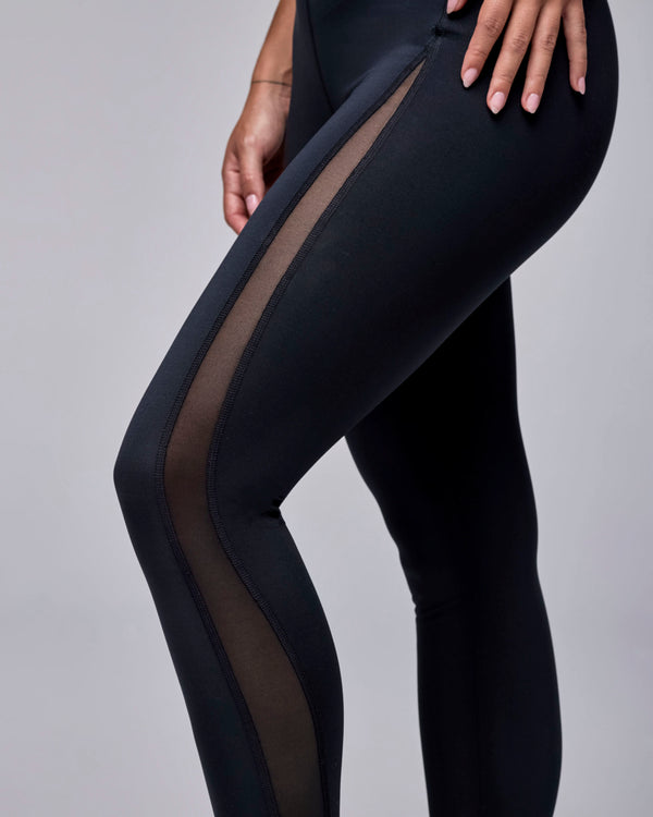 Close-up of the sheer black mesh insert curving from the hip down the outer leg of the black leggings, shown in side view.
