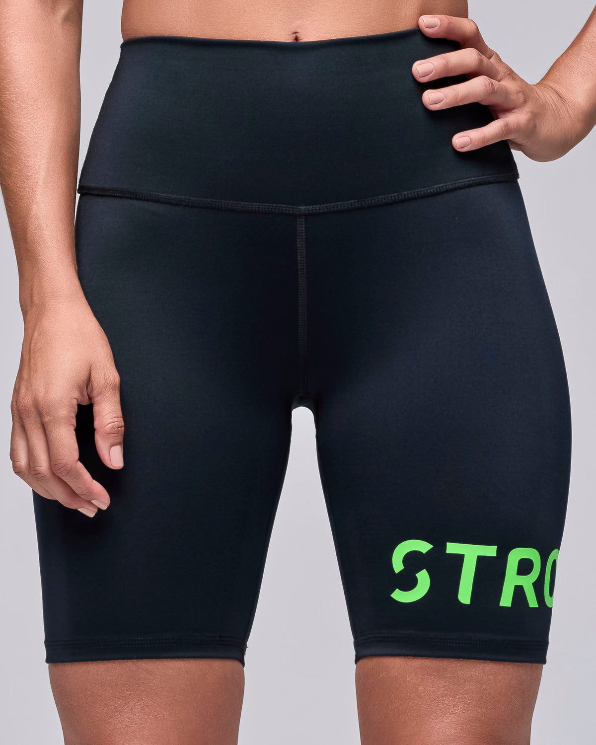 Female model in black high-waisted biker shorts with a wide, smooth waistband and large neon green 'STRONG iD' text wrapping around the left leg at the hem.