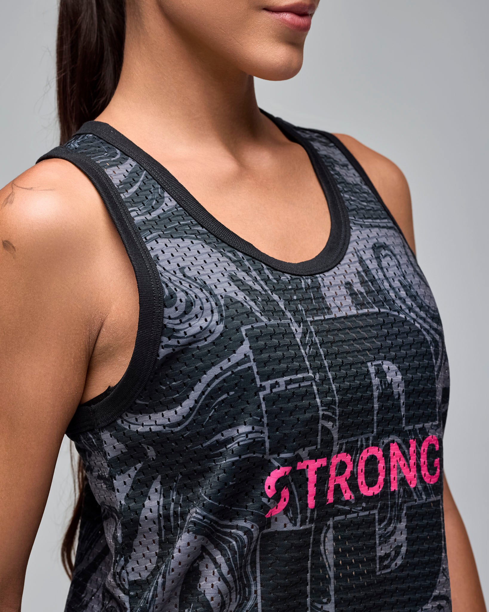 Close-up of a black and gray swirl-print mesh tank top with black trim and bright pink “STRONG iD” text on the front. Female model wears the sleeveless scoop-neck athletic tank against a light gray background.