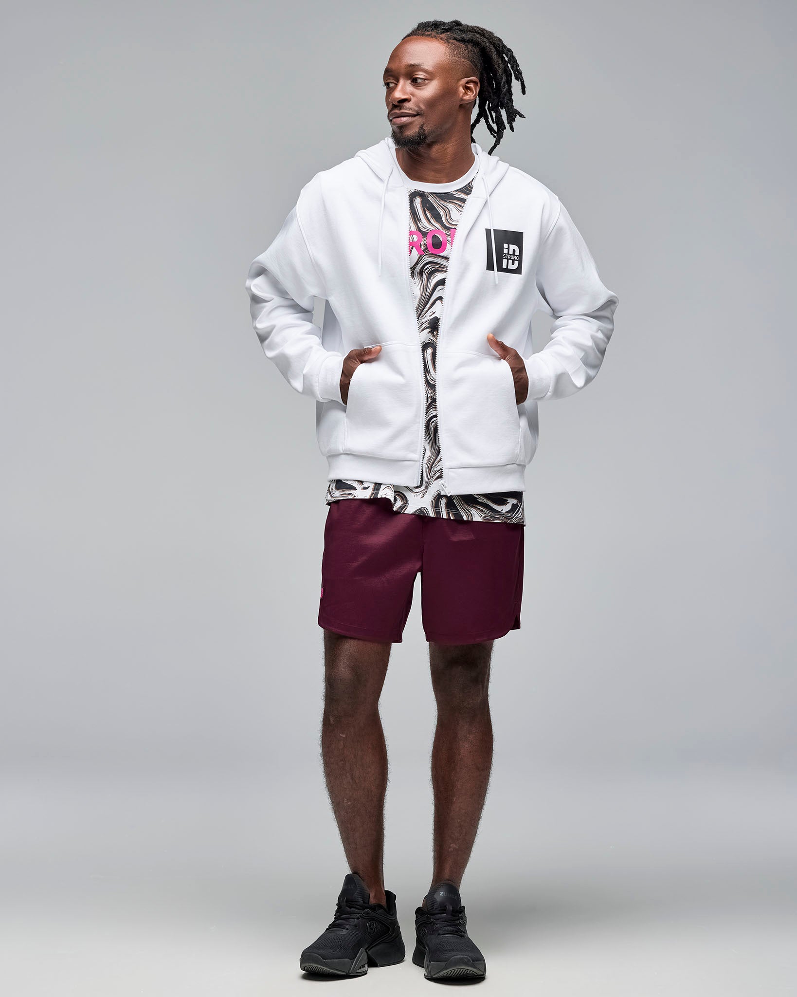 White zip-up hoodie with drawstrings and a black chest logo patch, layered over a white and black swirl-print graphic T-shirt with burgundy athletic shorts. Male model wears the hoodie unzipped with black sneakers, standing against a light gray background with hands in the front pockets.