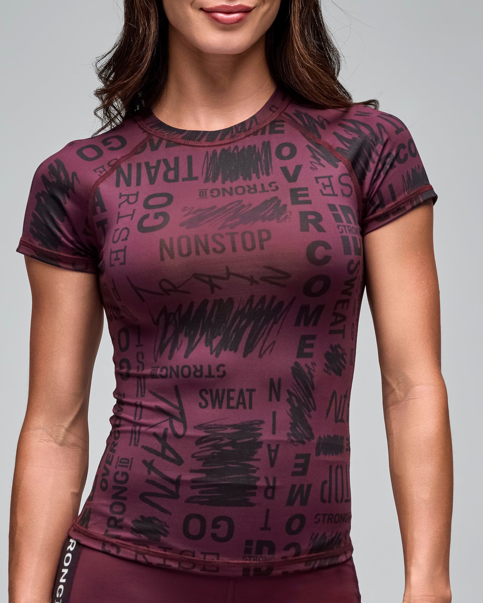 Burgundy fitted short-sleeve performance top with an allover black motivational word print and a crew neckline. Female model wears the sleek athletic tee with burgundy leggings in a close-up front view against a light gray background.
