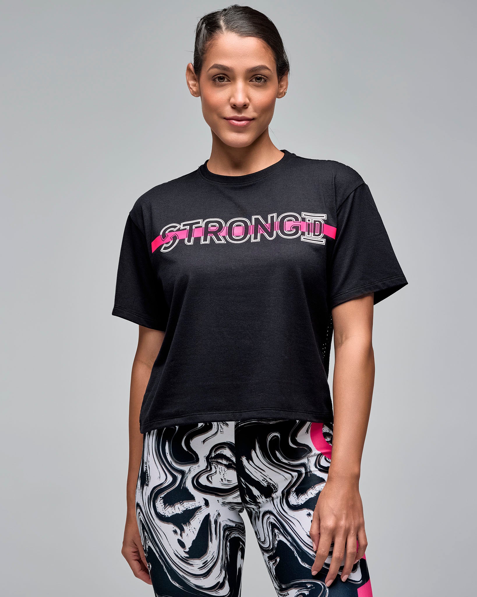 Black cropped short-sleeve T-shirt with white outlined “STRONG iD” text and a bright pink stripe across the chest. Female model wears the loose crewneck tee with white and black swirl-print leggings against a light gray background.