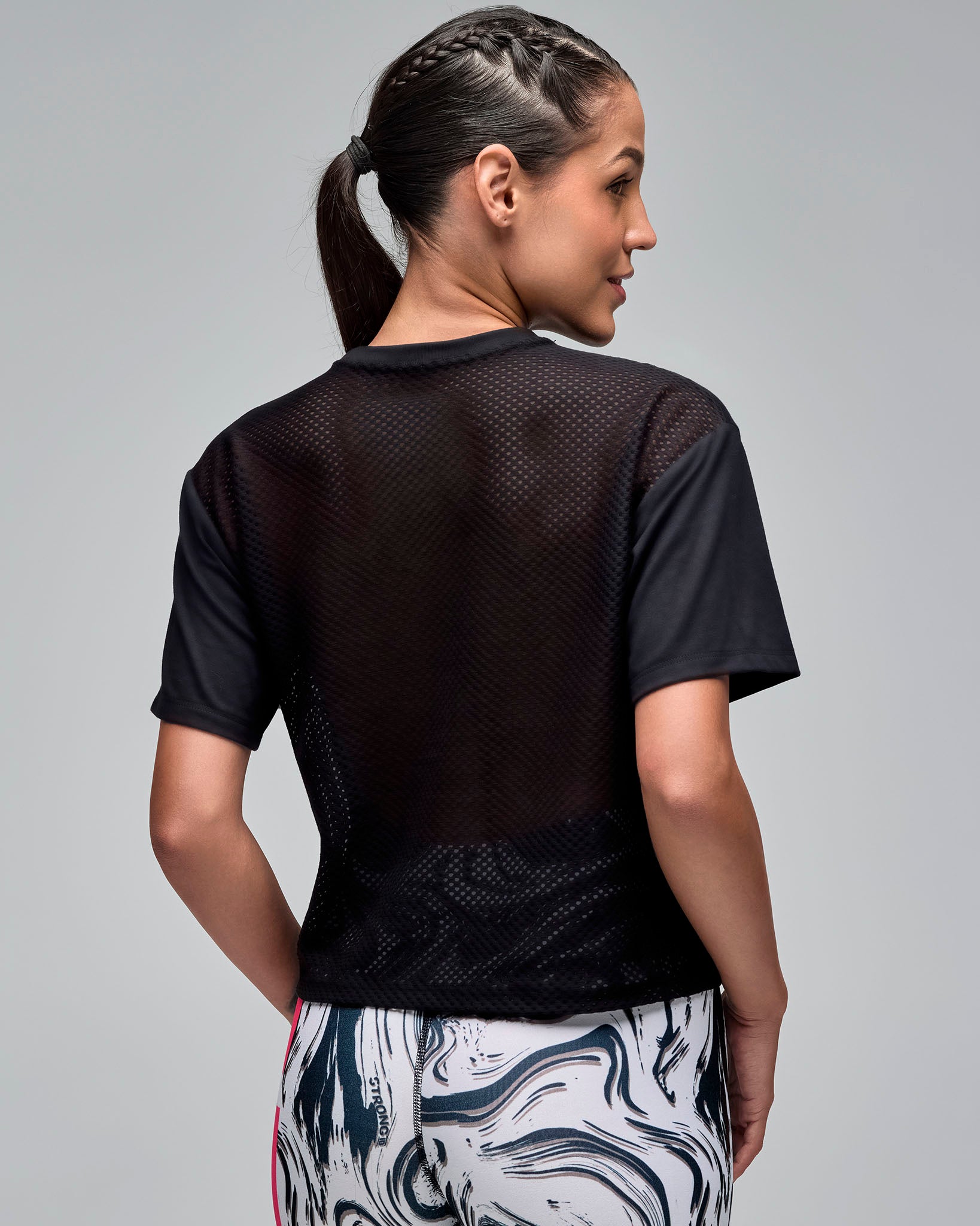 Back view of a black cropped short-sleeve T-shirt with a sheer mesh back panel. Female model wears the crewneck tee with white and black swirl-print leggings against a light gray background.