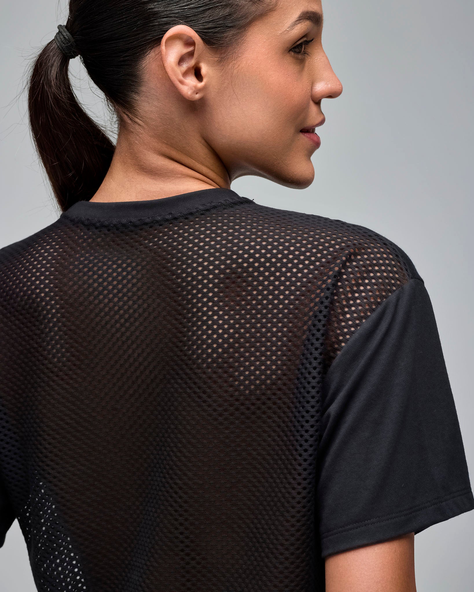 Close-up back view of a black cropped short-sleeve T-shirt with a sheer mesh back panel and solid black sleeves. Female model wears the tee against a light gray background.