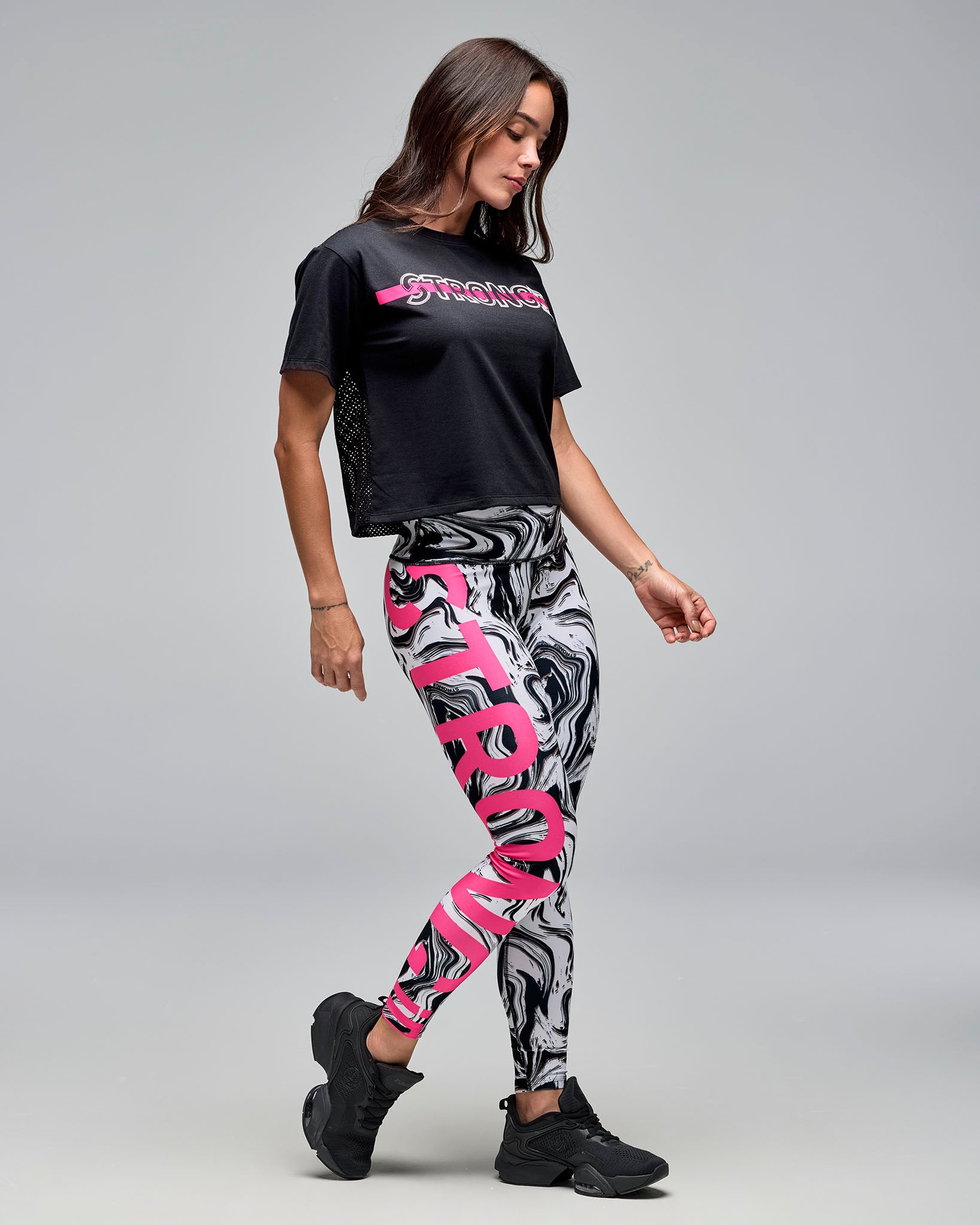 Black cropped short-sleeve T-shirt with a mesh back panel and the front has a white outlined “STRONG iD” text and a bright pink stripe across the chest. Female model wears the loose crewneck tee with white and black swirl-print leggings featuring oversized pink text and black sneakers against a light gray background.