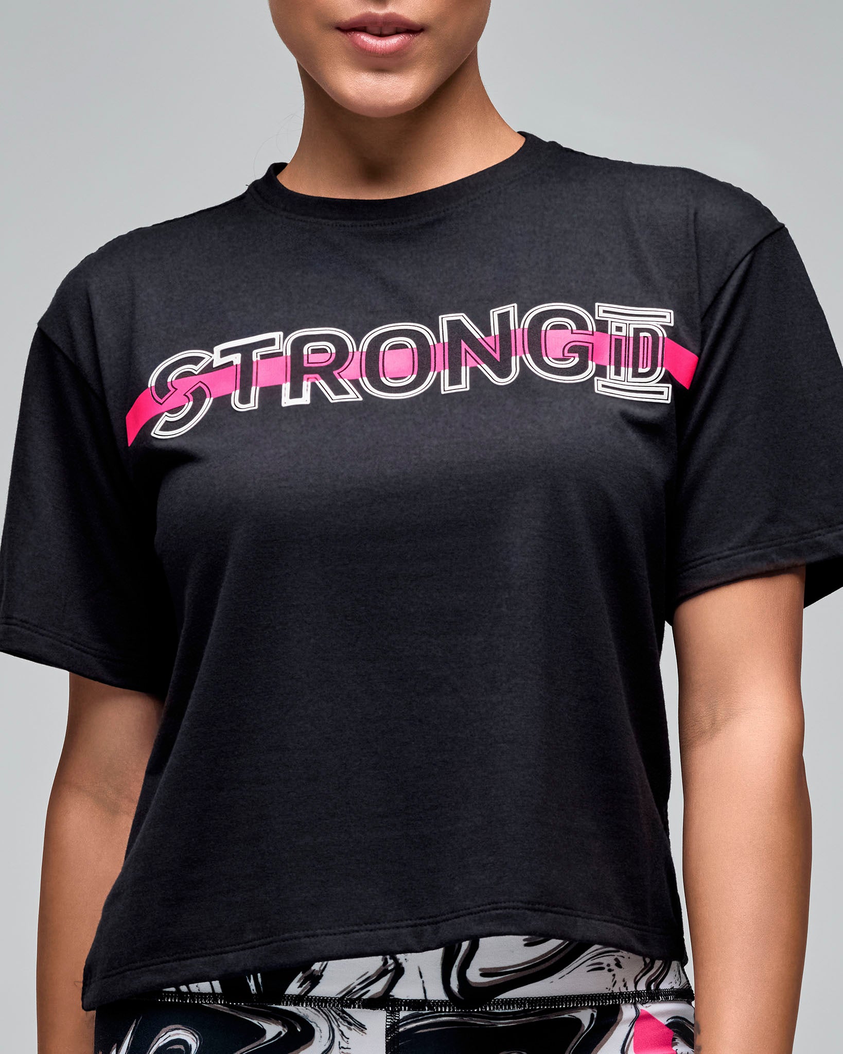 Close-up of a black cropped short-sleeve T-shirt with mesh back panel and a white outlined “STRONG iD” text and a bright pink stripe across the chest. Female model wears the loose crewneck tee over white and black swirl-print leggings against a light gray background.