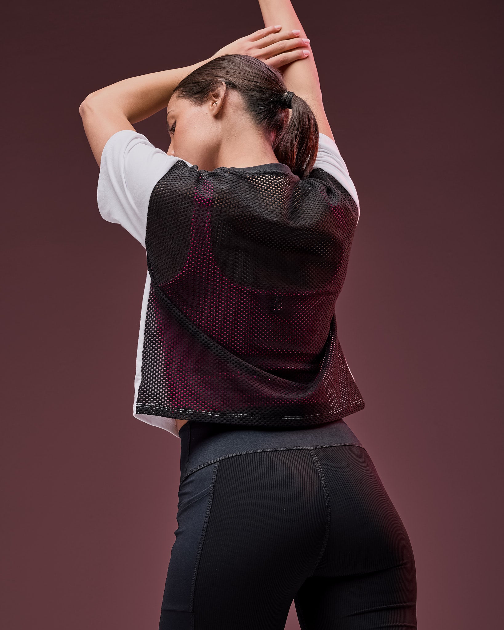 Black mesh short-sleeve athletic top layered over a white short-sleeve shirt, shown from the back on a female model wearing black high-waisted leggings against a dark mauve studio background.