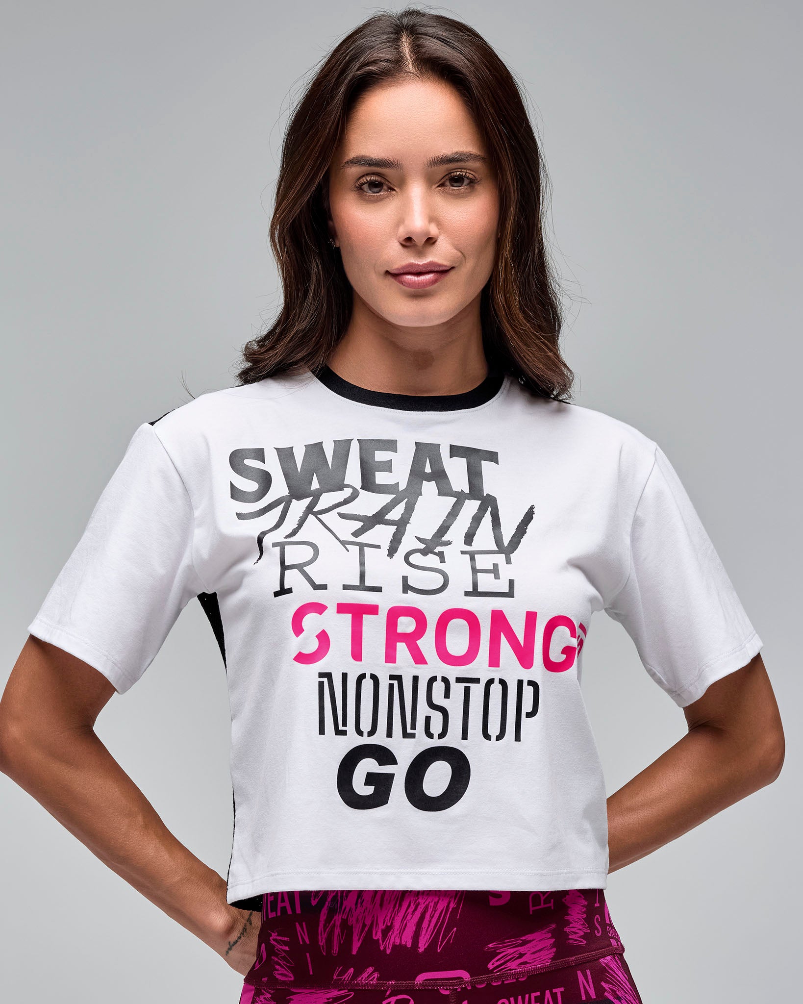 White cropped short-sleeve graphic T-shirt with black crew neckline, bold black “SWEAT TRAIN RISE,” bright pink “STRONG,” and black “NONSTOP GO” text on the front. Female model wears the boxy tee with bright pink patterned leggings against a light gray background.