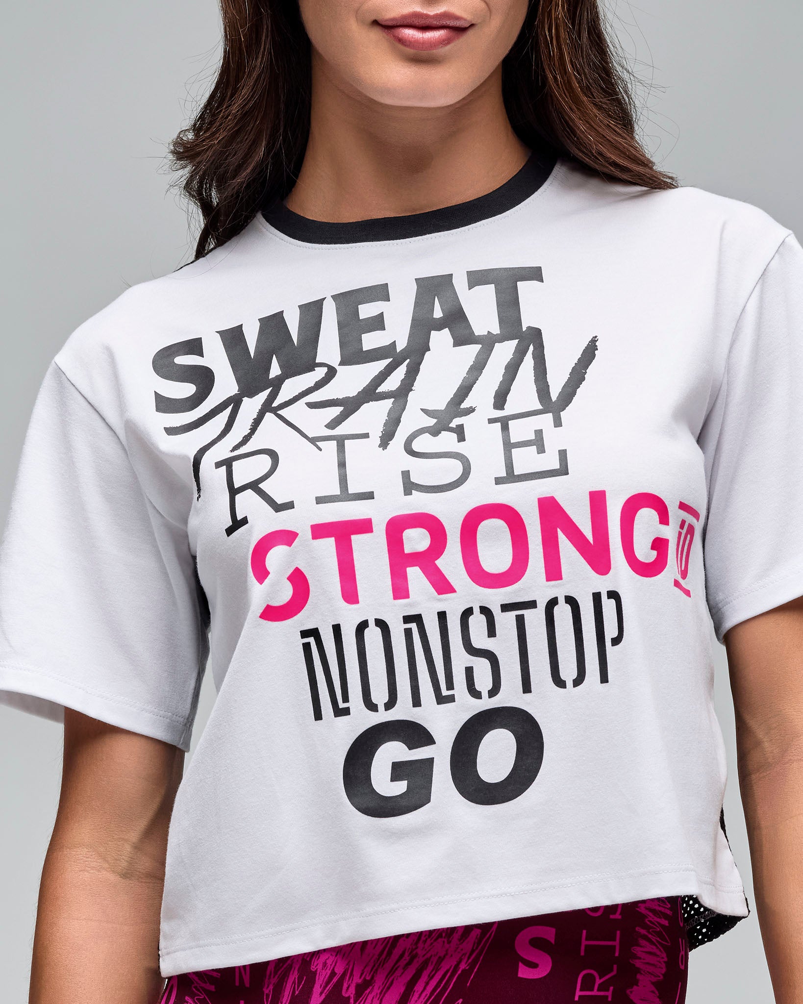 Close-up of a white cropped short-sleeve graphic T-shirt with black crew neckline and layered motivational text in black, gray, and bright pink across the front. Female model wears the tee with bright pink patterned leggings against a light gray background.