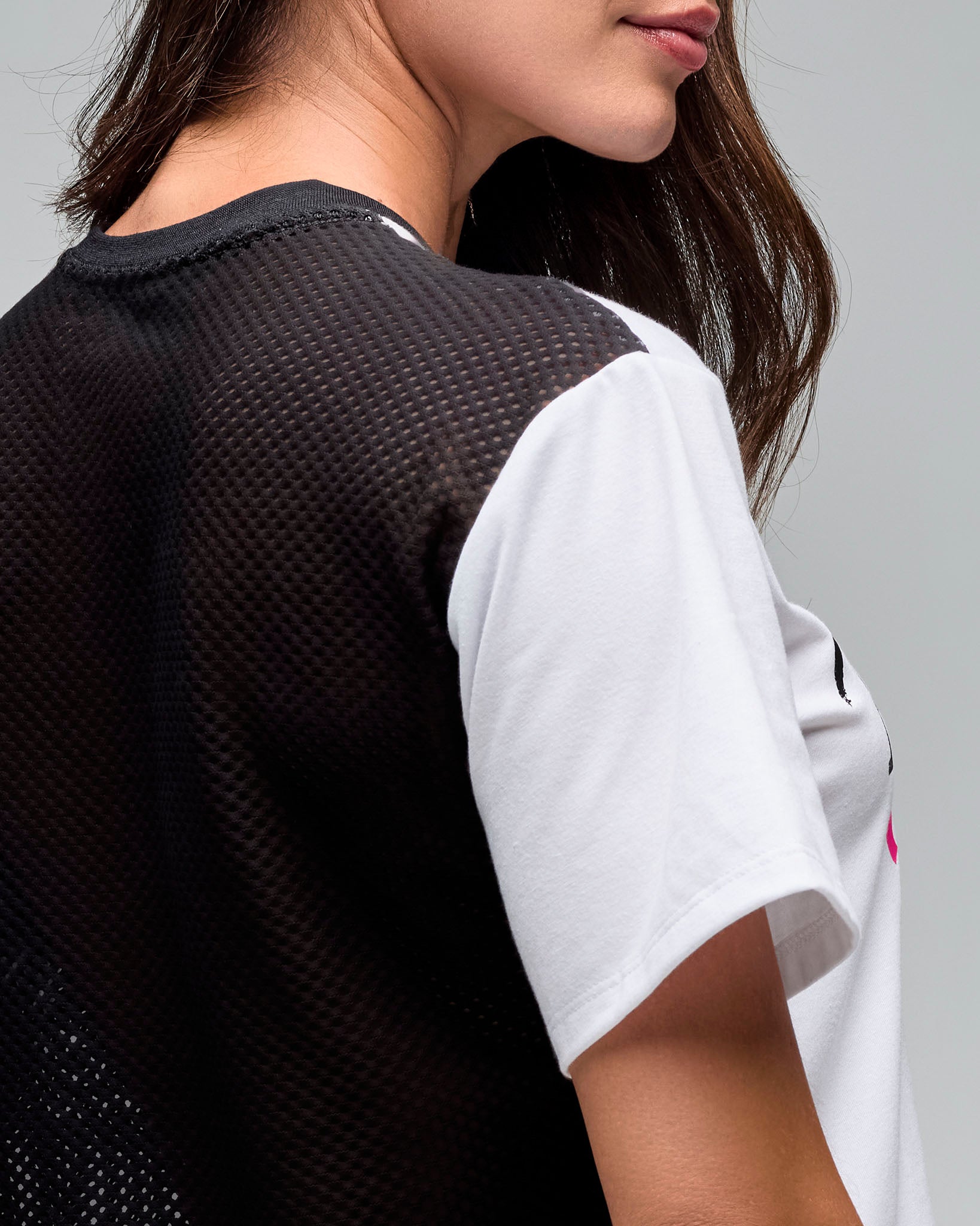 Back detail of a white cropped short-sleeve T-shirt with a black mesh back panel and white sleeves. Female model wears the tee against a light gray background.