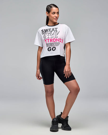 White cropped short-sleeve graphic T-shirt with black crew neckline, black mesh back panel, and black, gray, and bright pink motivational text on the front. Female model wears the tee with black biker shorts and black sneakers against a light gray background.