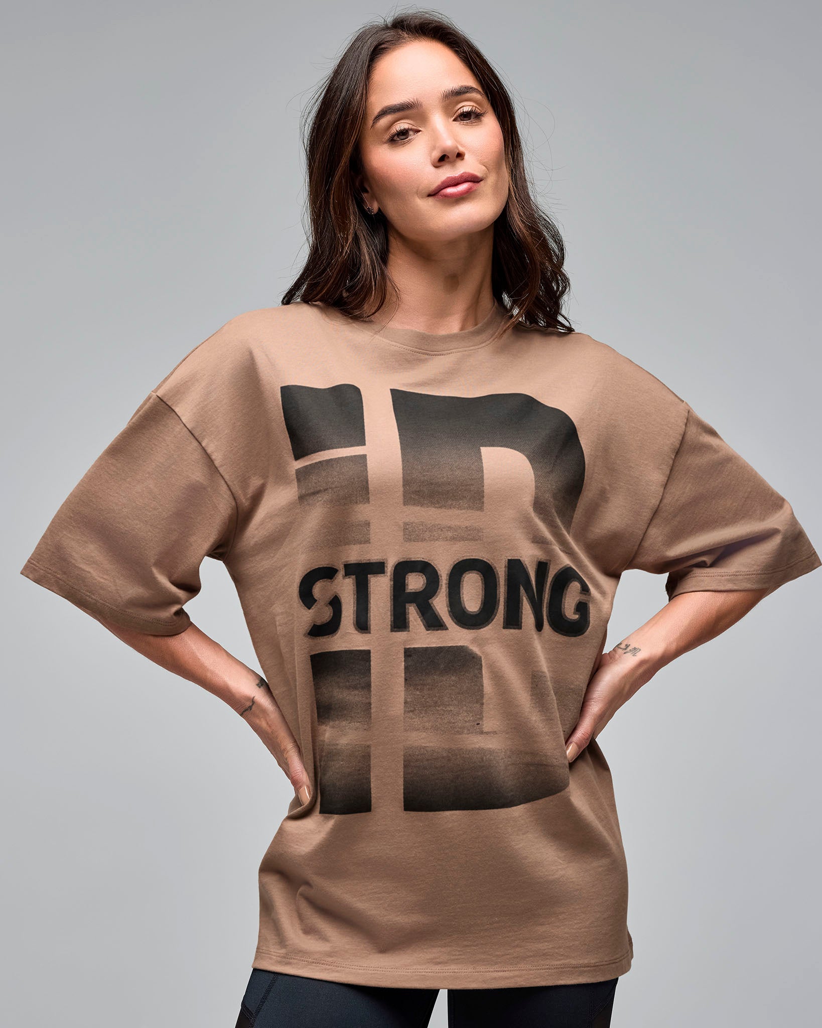 Taupe oversized graphic T-shirt with a large black "STRONG iD" front print and crew neckline, shown on a female model wearing black leggings against a light gray studio background.