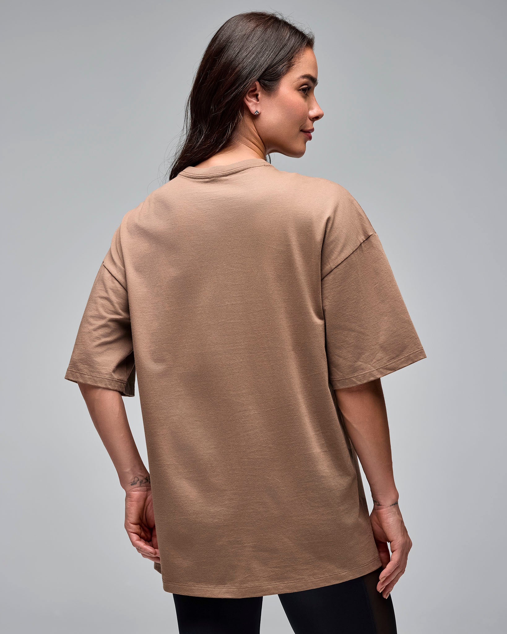 Taupe oversized short-sleeve T-shirt with a plain back and crew neckline, shown from the back on a female model wearing black leggings against a light gray studio background.