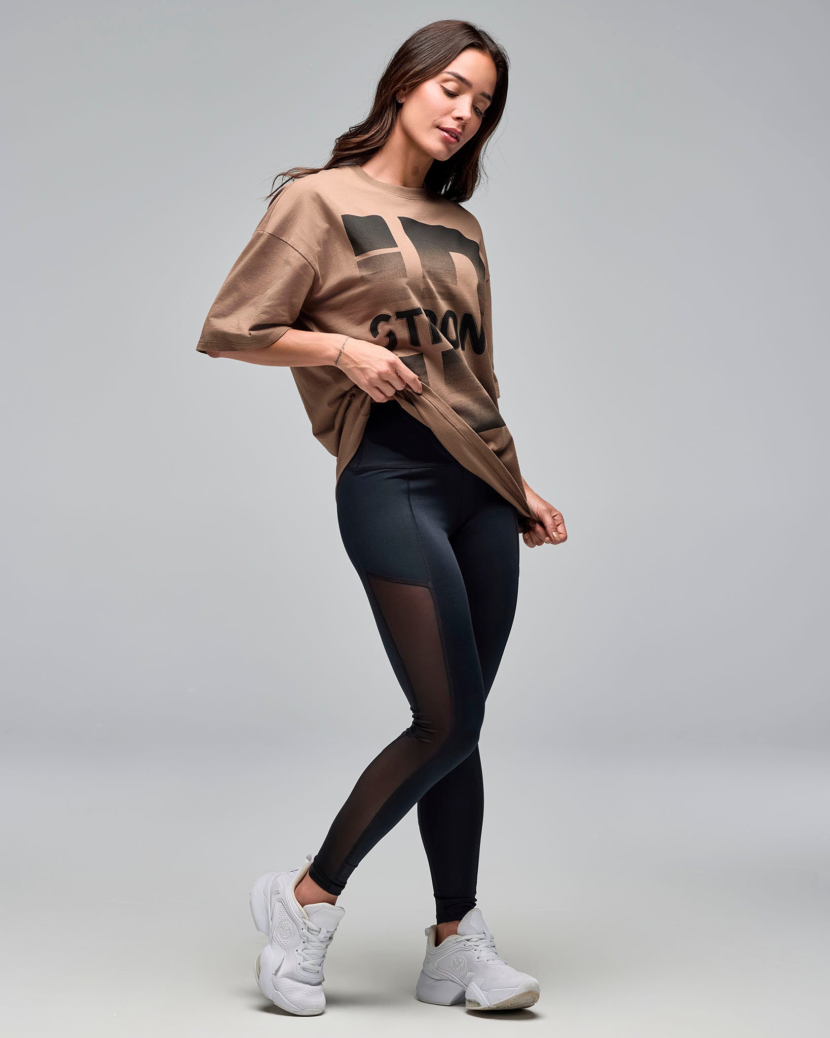 Taupe oversized graphic T-shirt with large black “STRONG ID” graphic print, short sleeves and a crew neckline, shown on a female model wearing black high-waisted leggings with sheer side panels and white sneakers against a light gray studio background.