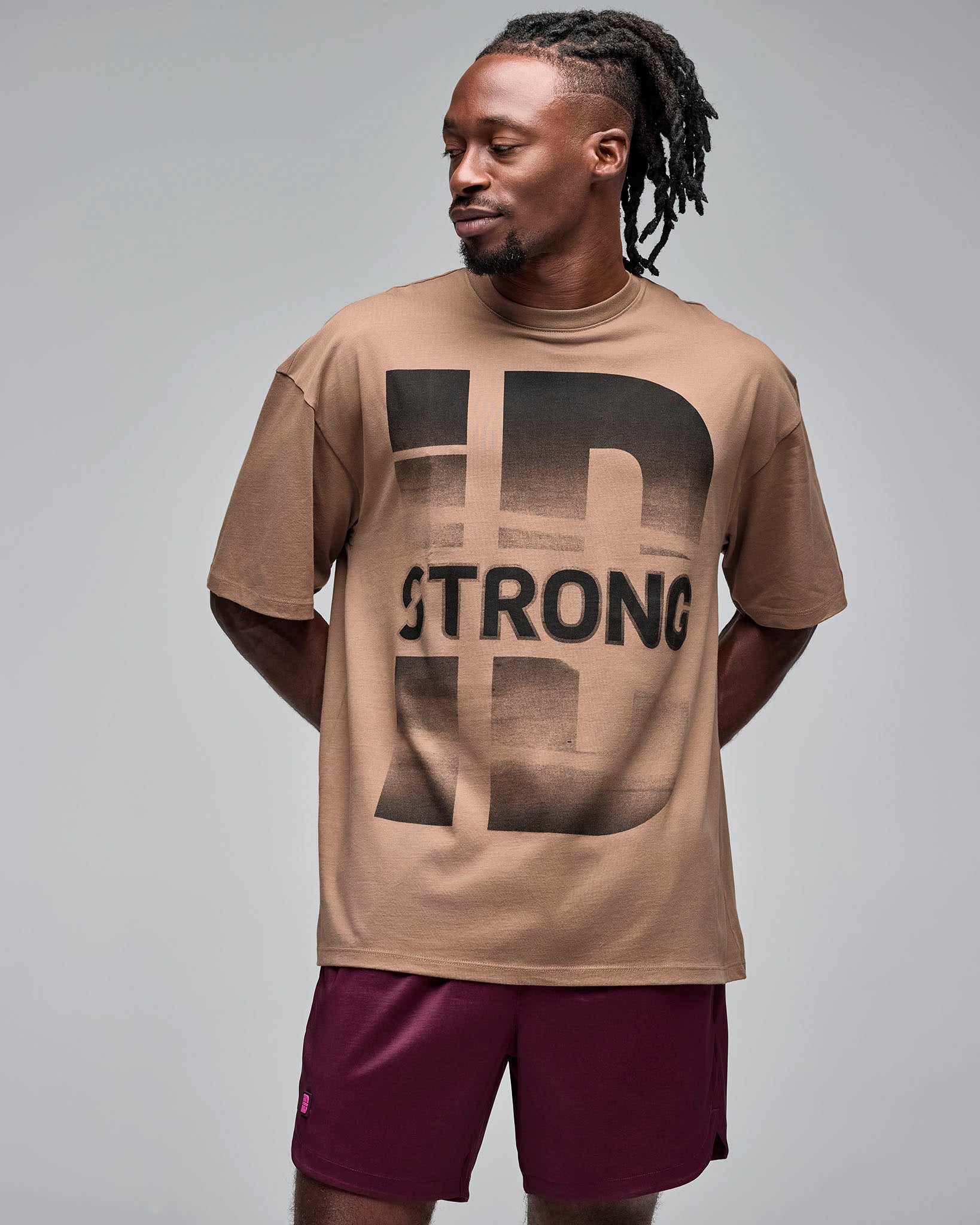 Taupe oversized T-shirt with a large black “STRONG ID” graphic print and crew neckline, shown on a male model wearing burgundy athletic shorts against a light gray studio background.