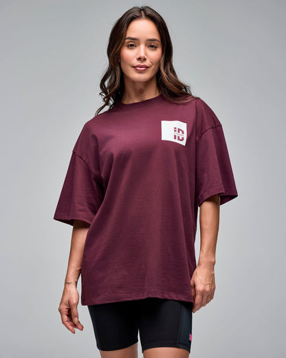 Burgundy oversized short-sleeve T-shirt with a small white chest logo patch. Female model wears the loose-fit tee over black biker shorts against a light gray background.