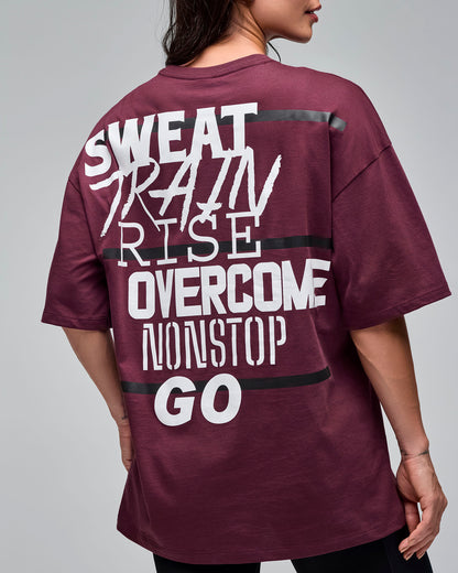 Back view of a burgundy oversized short-sleeve T-shirt with large white “SWEAT TRAIN RISE OVERCOME NONSTOP GO” text and black stripe details across the back. Female model wears the tee with black biker shorts against a light gray background.