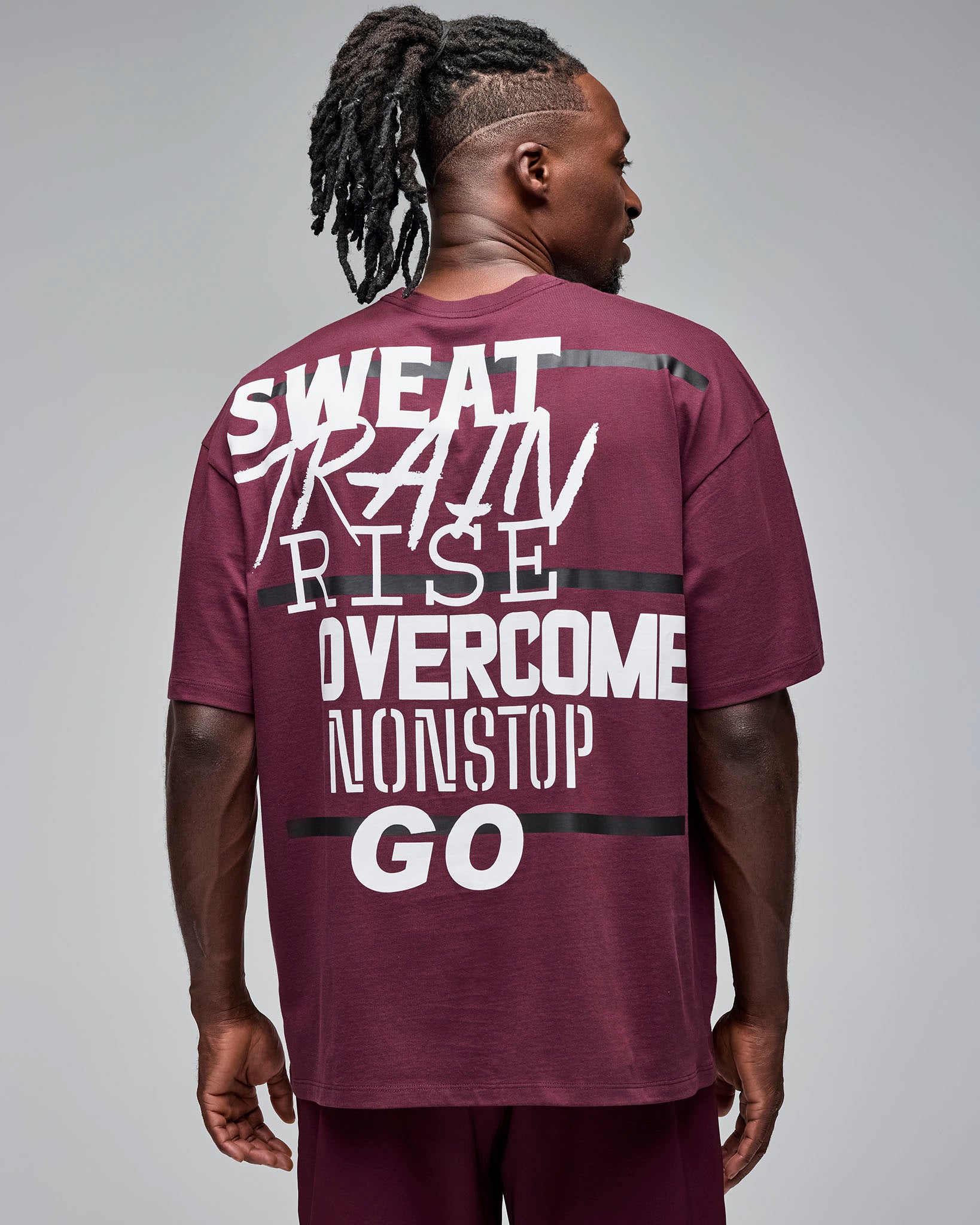 Back view of a burgundy oversized short-sleeve T-shirt with large white motivational text and black stripe accents across the back. Male model wears the tee with burgundy shorts against a light gray background.