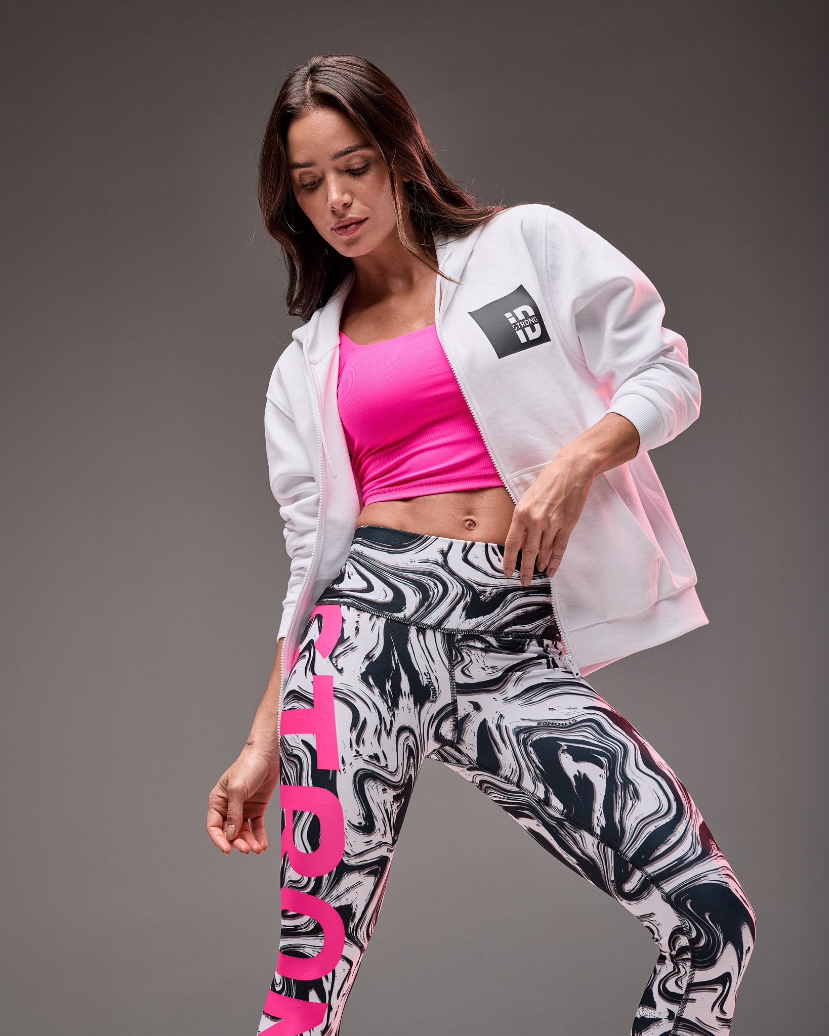 White zip-up hoodie with a small black and white "STRONG iD" chest graphic, layered over a bright pink cropped athletic top and paired with black, white, and gray marble-print leggings with bold pink lettering on a female model.