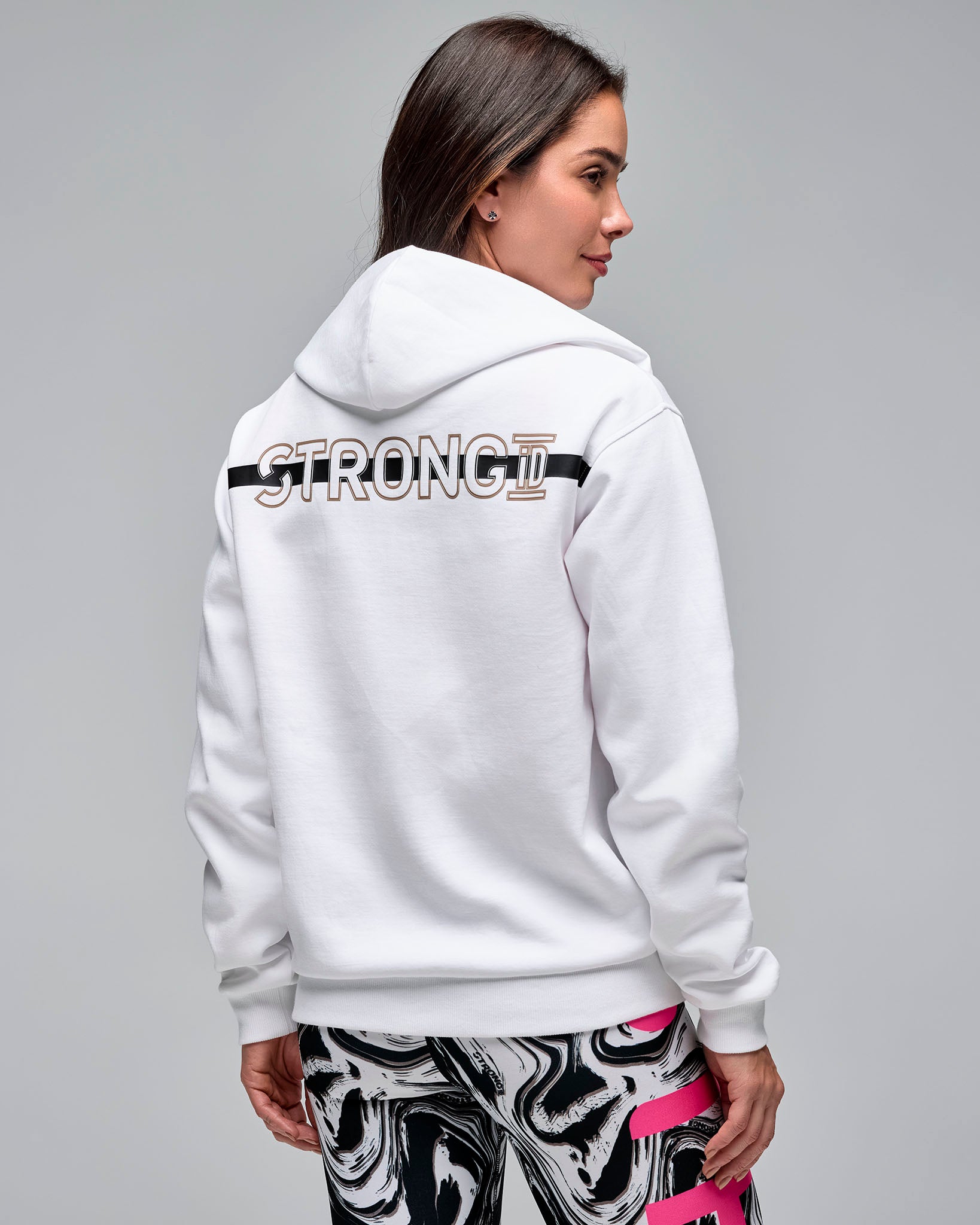 Back view of a white zip-up hoodie with a hood and large “STRONG iD” graphic across the upper back. Female model wears the long-sleeve hoodie over white and black swirl-print leggings against a light gray background.