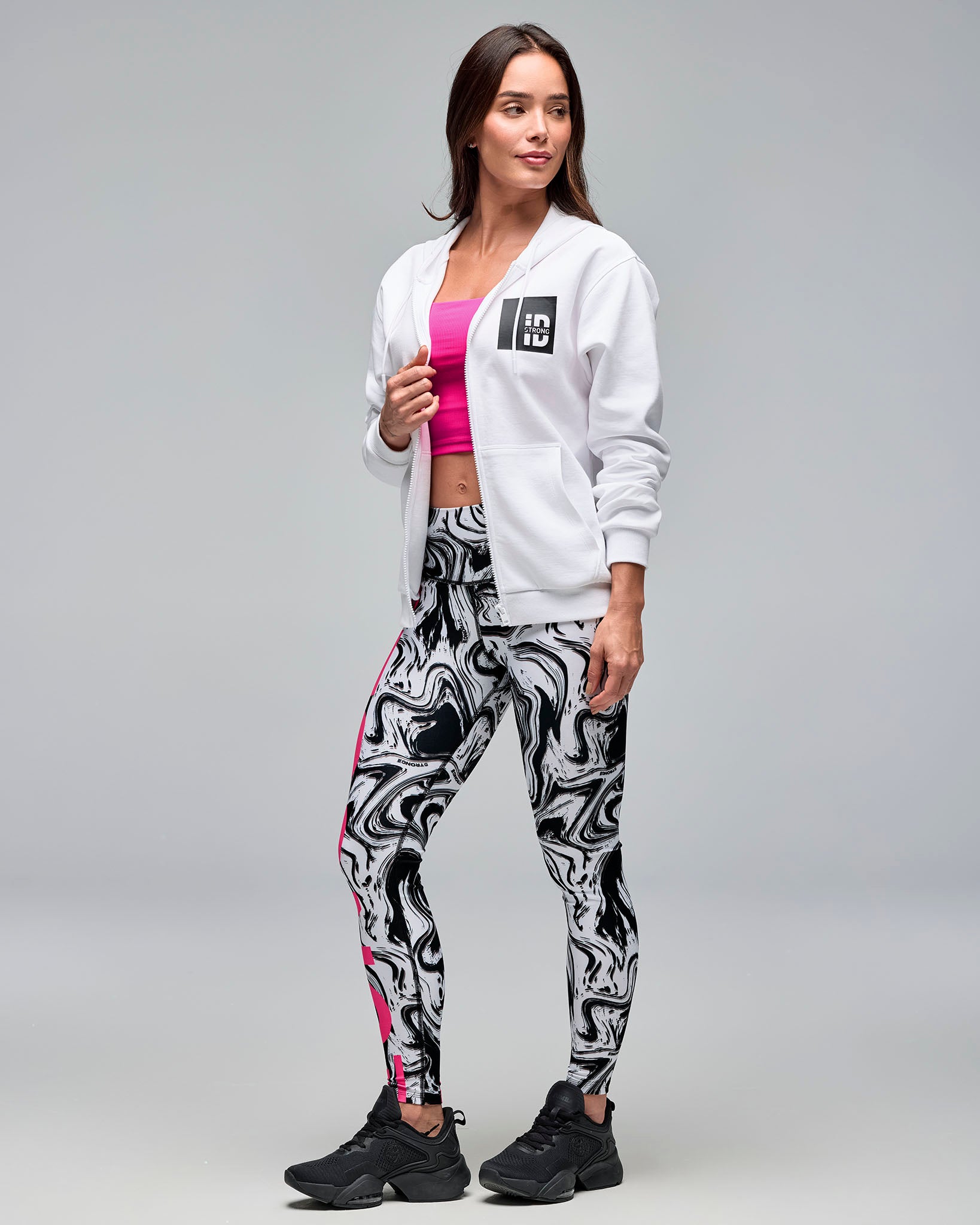White zip-up hoodie with drawstrings, front pockets, and a black chest logo patch. Female model wears the long-sleeve hoodie open over a bright pink crop top, white and black swirl-print leggings, and black sneakers against a light gray background.
