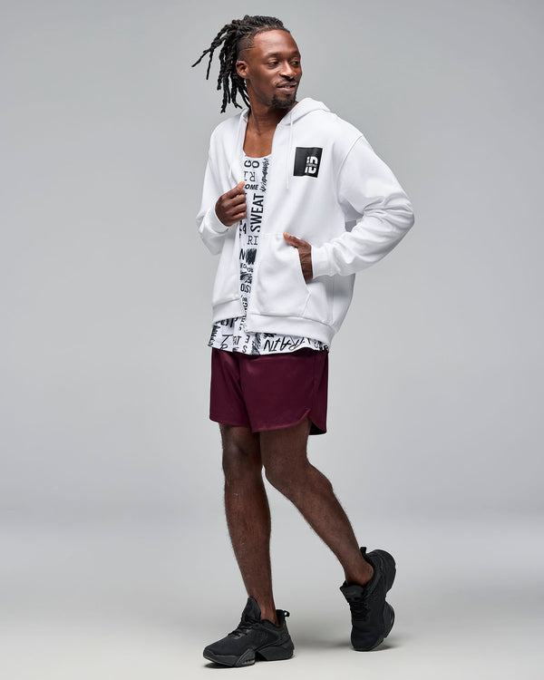White zip-up hoodie with drawstrings, front pockets, and a black chest logo patch. Male model wears the long-sleeve hoodie open over a white graphic tank, burgundy athletic shorts, and black sneakers against a light gray background.