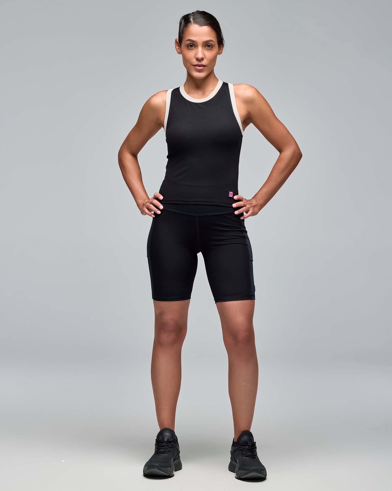 Black sleeveless athletic tank with white trim at the neckline and armholes, shown on a female model with black fitted biker shorts and black sneakers against a light gray studio background.