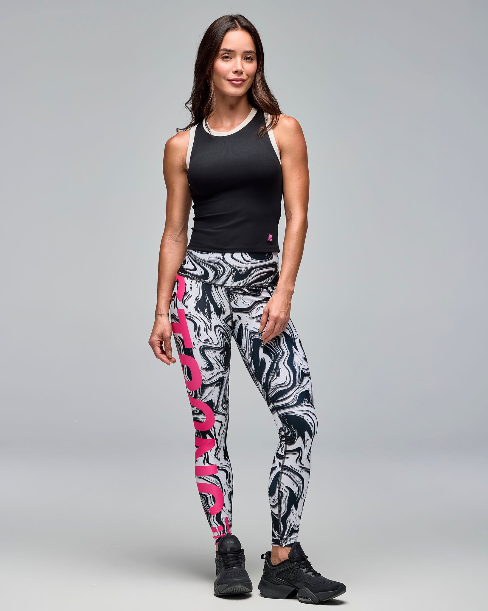 Black sleeveless athletic tank with white trim at the neckline and armholes, shown full length on a female model wearing high-waisted black, white, and gray marble-print leggings with bold pink lettering and black sneakers.