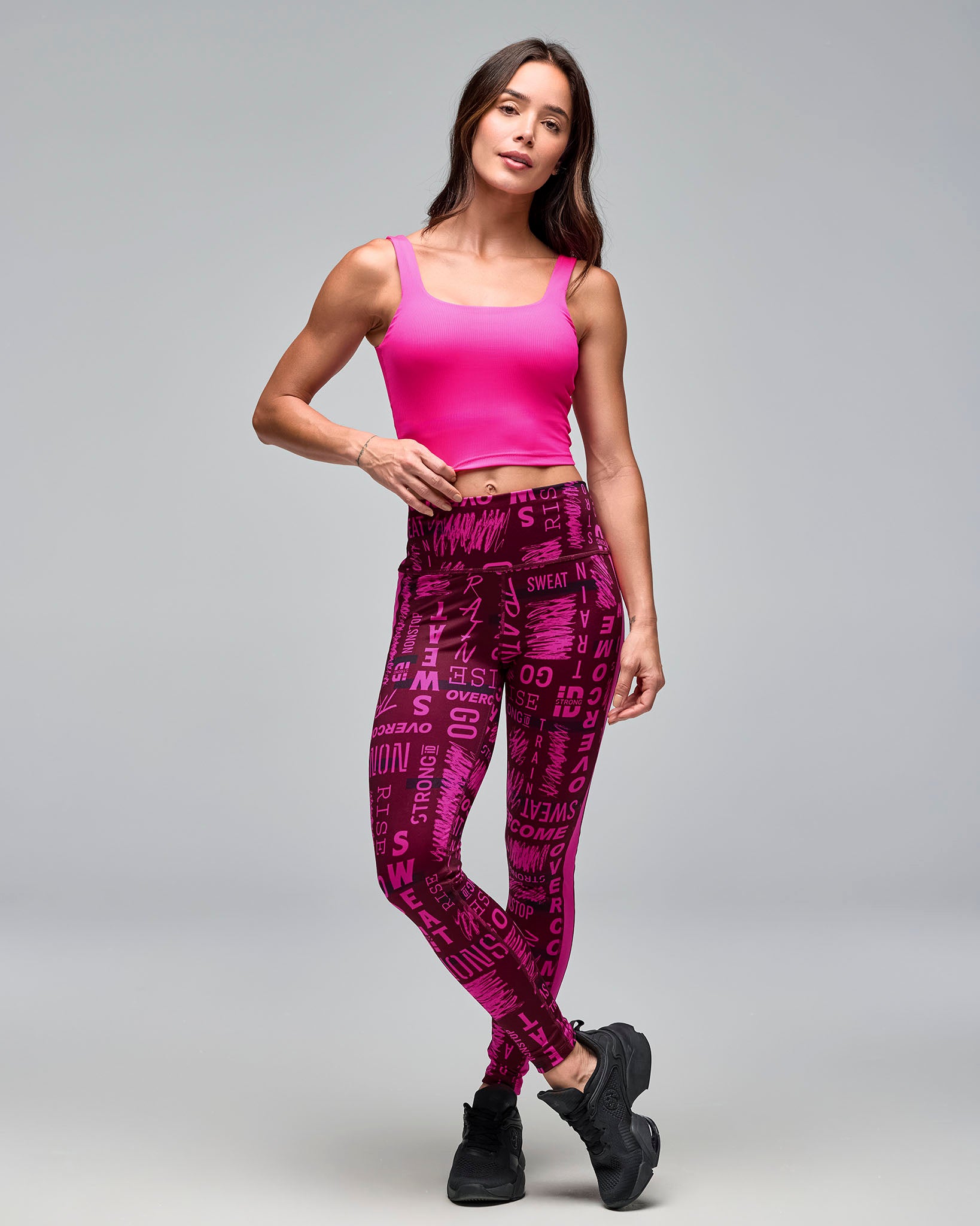 Pink cropped athletic tank with wide shoulder straps and a square neckline, shown full length on a female model wearing burgundy and pink graphic-print high-waisted leggings and black sneakers against a light gray studio background.