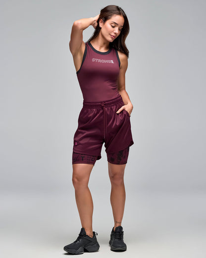 Burgundy fitted racerback tank top with black trim and white “STRONG iD” text on the chest. Female model wears the sleeveless athletic top with burgundy athletic shorts layered over burgundy printed biker shorts and black sneakers against a light gray background.