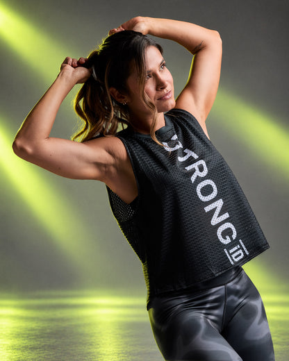 Female model in a loose black open-hole mesh muscle tank with vertical white 'STRONG iD' text down the front, paired with black and grey swirl print leggings, against neon light beams.
