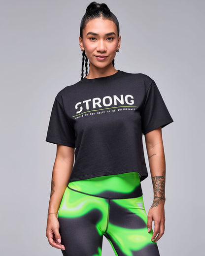 Female model in a black cropped boxy top with white 'STRONG' text, a neon green line, and the small tagline 'UNITED IN OUR QUEST TO BE UNSTOPPABLE' across the chest. Paired with neon green and black swirl print leggings.