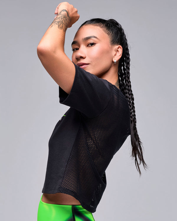 Female model with one arm raised in the black 'STRONG' boxy top, shown in side view with the mesh back and short sleeves.